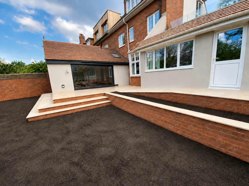 Completed external works at a large property: pale paved terraces, brick retaining walls and wide steps stepping down to prepared ground, with a rendered extension beyond.