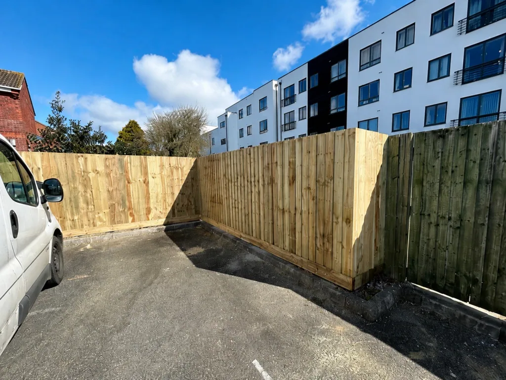 Newly installed close-board timber fencing returning around the corner of a tarmac parking area, with apartment buildings beyond.
